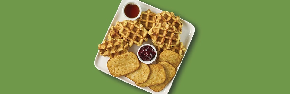 White platter with hashbrowns and waffles and dipping bowls of maple syrup and cranberry sauce. Platter is on a green back-ground.