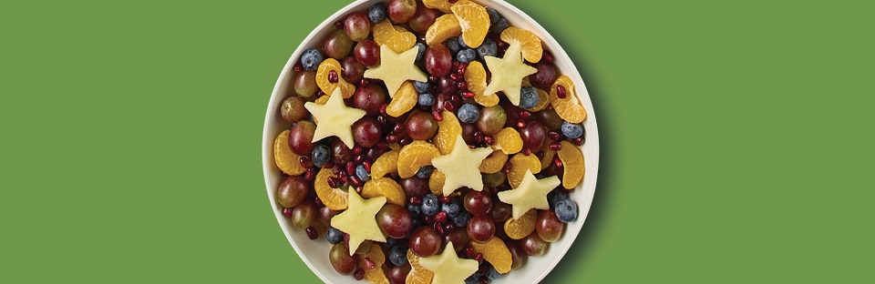 Fruit salad in white bowl. Salad has grapes, blueberries, orange segments, pome-granate arils and apples cut into star shapes. Salad is on a green background.
