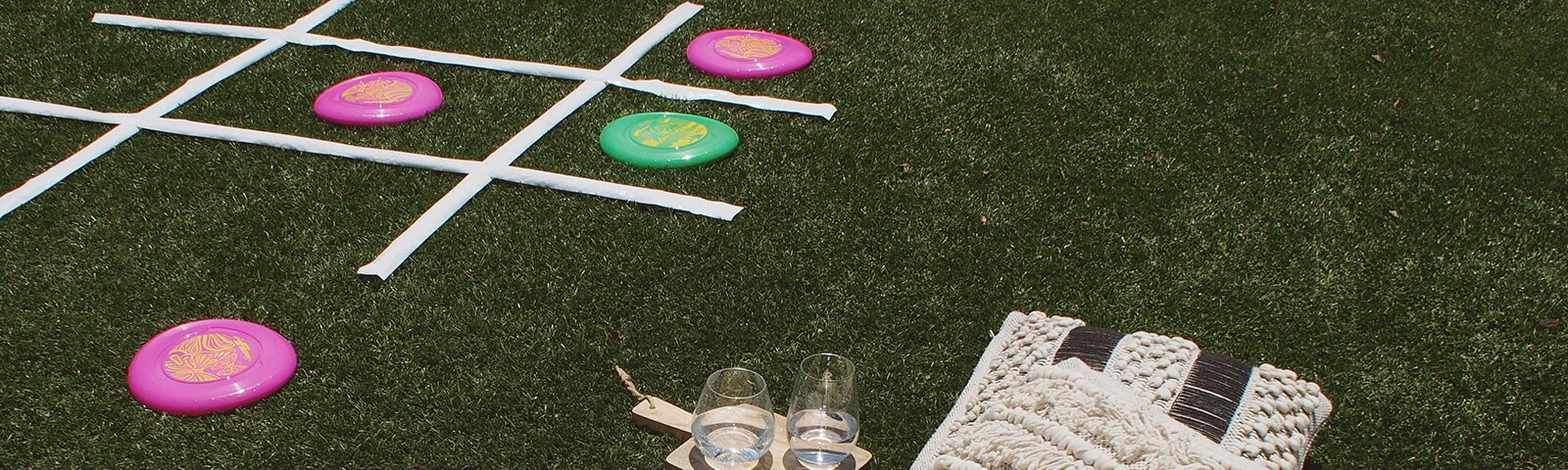 A large tic-tac-toe board sits on an artificial field. Frisbees are being used to mark the players and a blanket and pillows are setup nearby A large tic-tac-toe board sits on an artificial field. Frisbees are being used to mark the players and a blanket and pillows are setup nearby