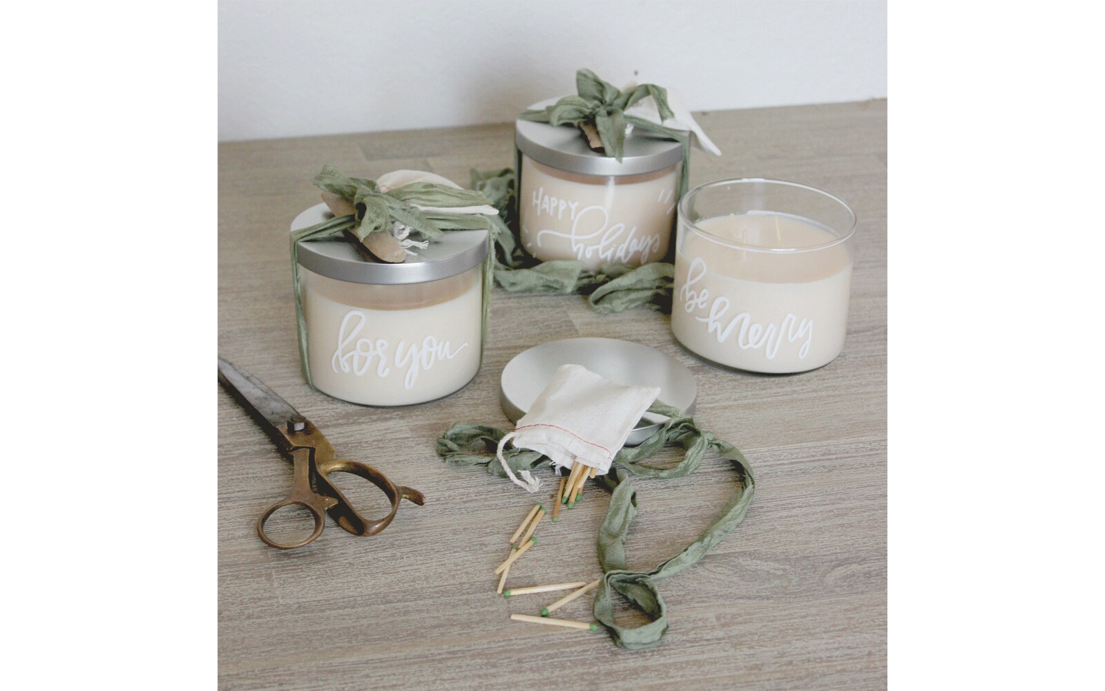 White candles with silver lids and words written on the glass sit on a table next to scissors and matches White candles with silver lids and words written on the glass sit on a table next to scissors and matches