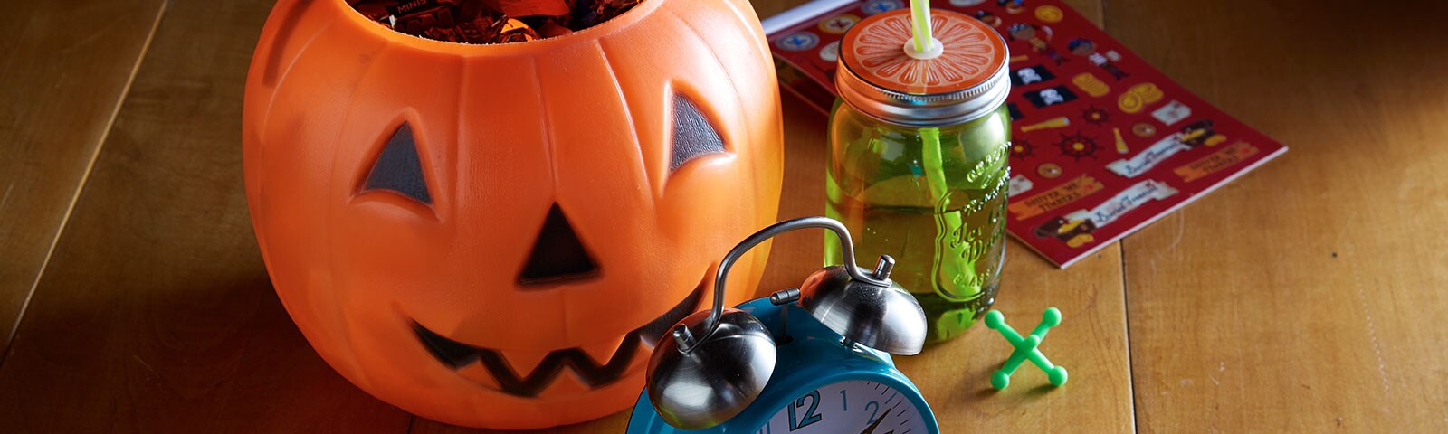 A jack-o-lantern candy bucket, a mason jar with lid and straw, and an alarm clock sitting on a wooden table A jack-o-lantern candy bucket, a mason jar with lid and straw, and an alarm clock sitting on a wooden table