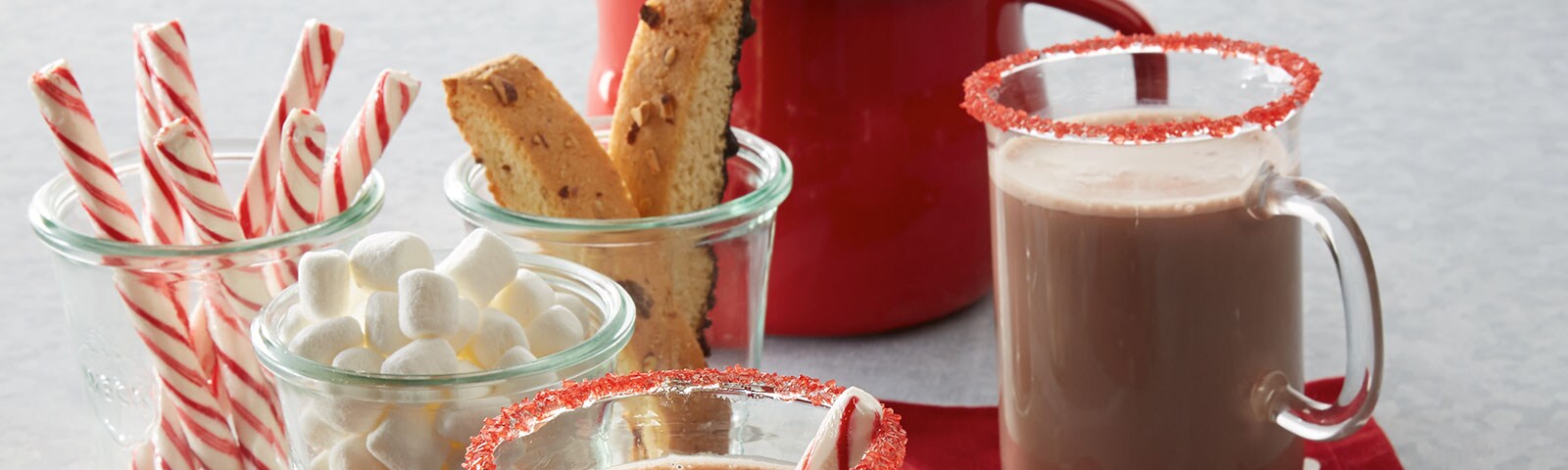 A cup of hot cocoa with a colorful red candied rim sits next to three cups containing candy cane sticks, biscotti and marshmallow respectively A cup of hot cocoa with a colorful red candied rim sits next to three cups containing candy cane sticks, biscotti and marshmallow respectively