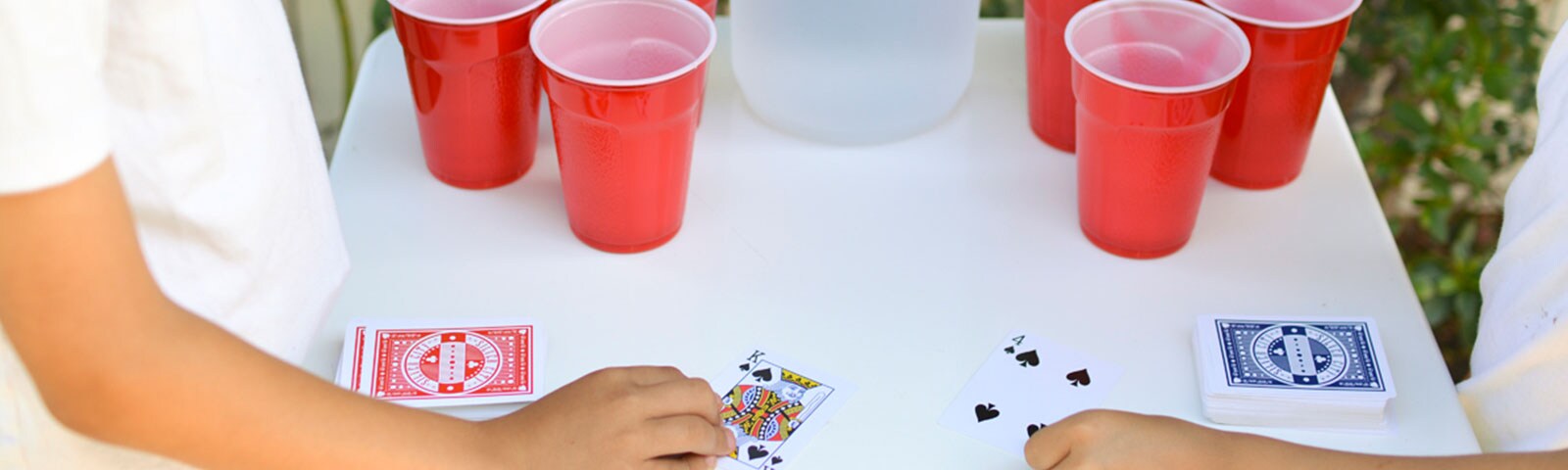 On an outdoor table 2 sets of 4 red solo cups sit with a jug of water in the middle while 2 people stand on each side facing off in a card game. On an outdoor table 2 sets of 4 red solo cups sit with a jug of water in the middle while 2 people stand on each side facing off in a card game.